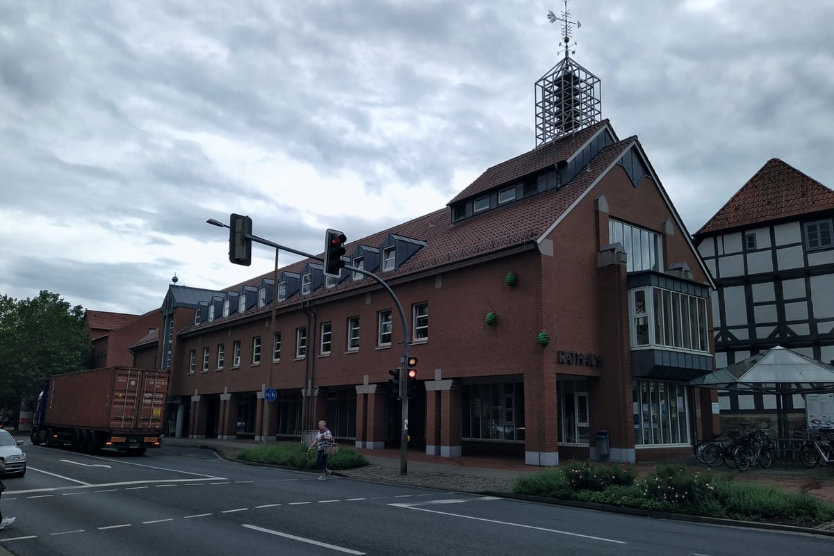 Rathaus Wunstorf: roter Backsteinbau mit Giebel und stählernem Turmaufsatz an einer Straßenkreuzung, rechts ein Fachwerkhaus der Altstadt