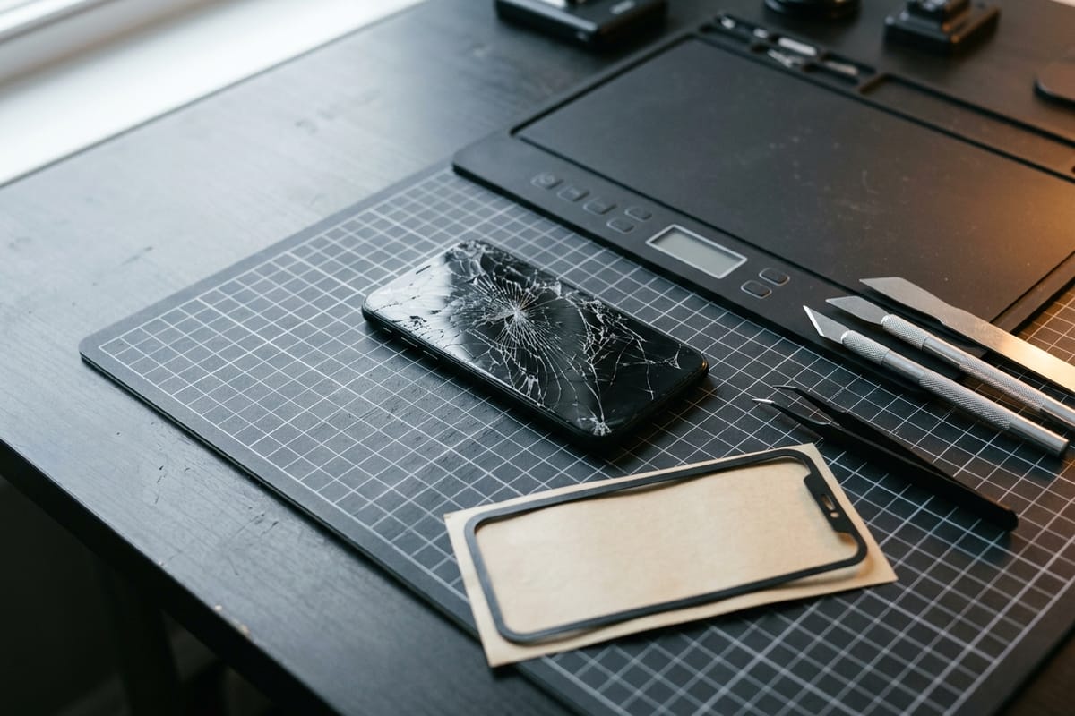 Die gesprungene Glasrückseite eines Smartphones liegt neben einer Heizmatte und dünnen Öffnungsklingen.